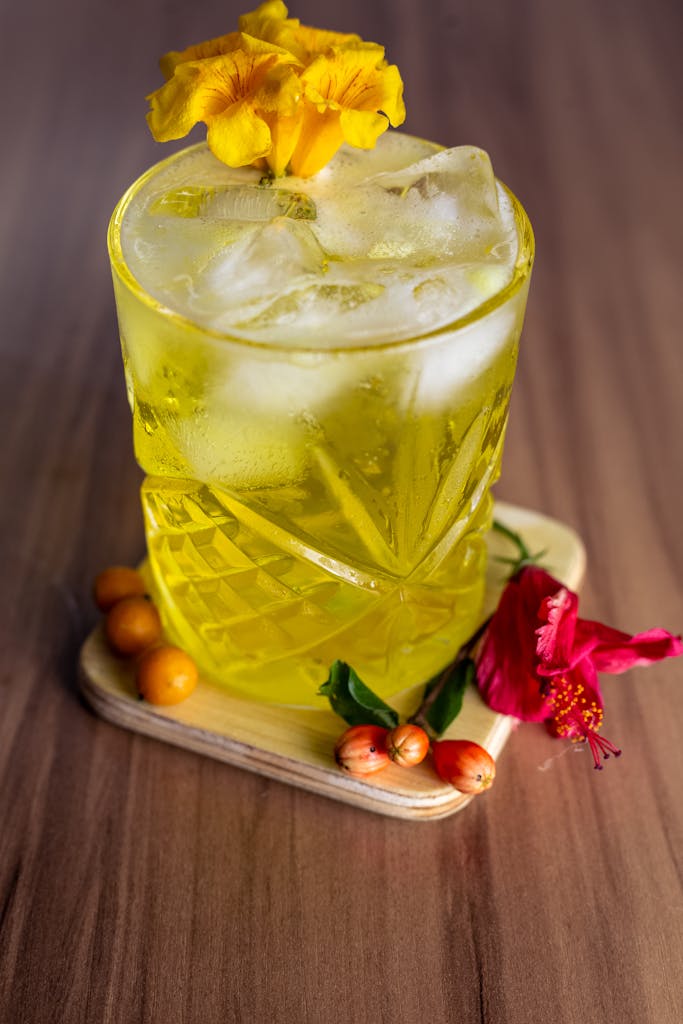 Bright yellow drink with ice cubes and floral garnish on a wooden table.