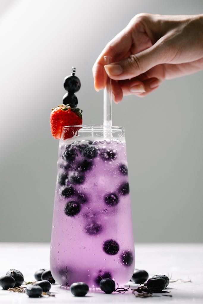 Close-up of a hand holding a glass of sparkling blueberry drink with a strawberry garnish.