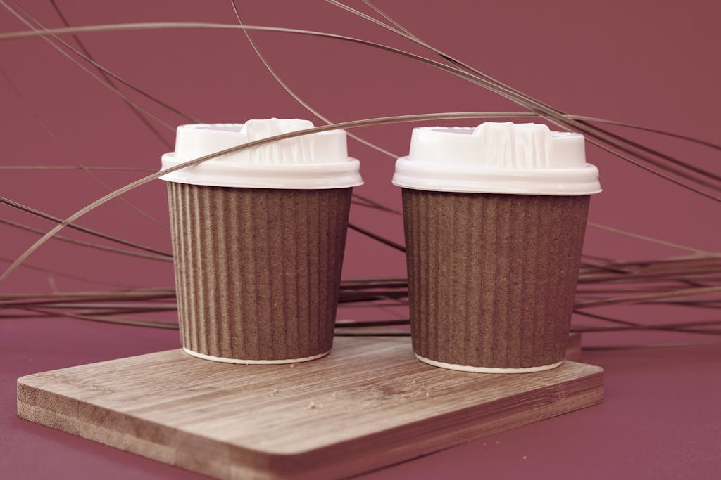 Two eco-friendly disposable coffee cups with lids on a wooden board, set against a pink background.