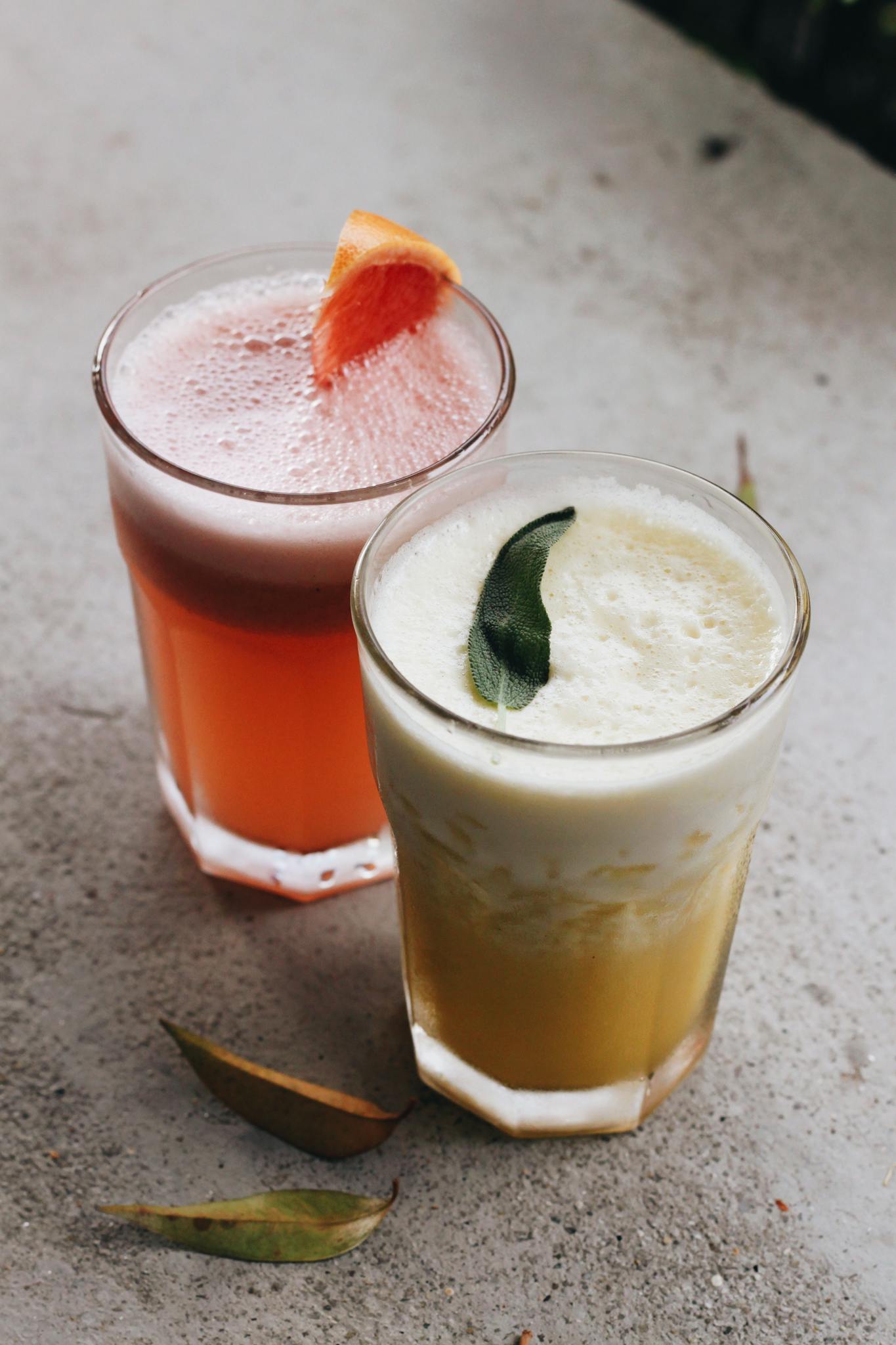 Two vibrant citrus and tropical fruit drinks in glasses on a textured table, garnished with fruit slices and leaves.